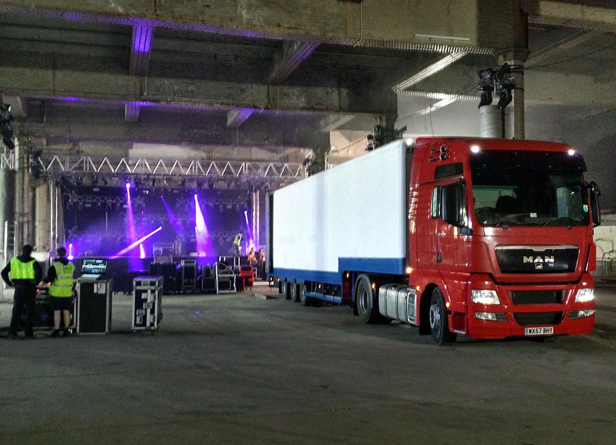 Red artic with a white trailer in front of a stage with purple lighting, with two crew in hi-vis jackets